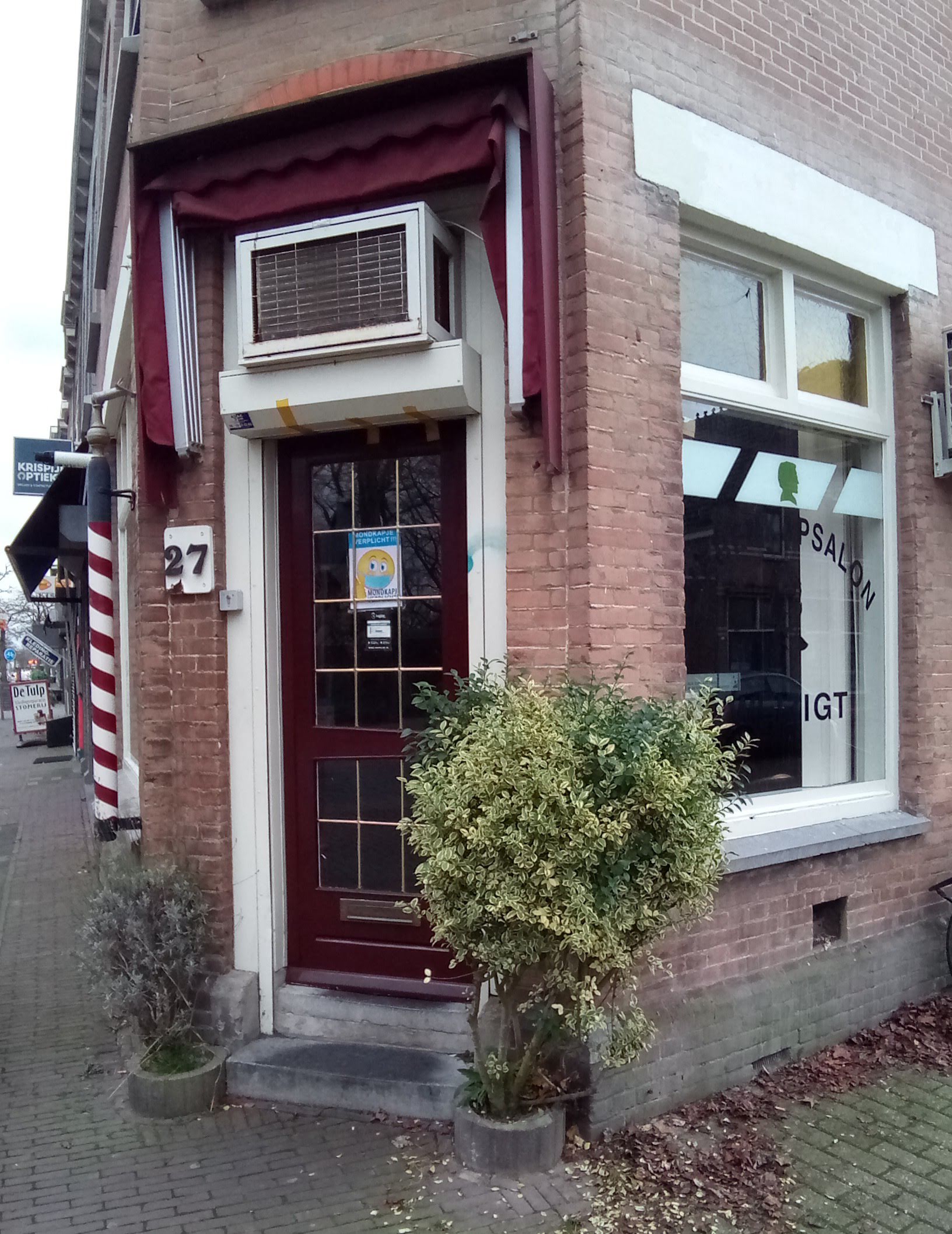 De hoekpand-gevel van Herenkapsalon Weizigt: bordeauxrode deur met nummer 27, luifel en een rood-witte kapperspaal